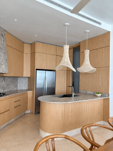 Vista general de la cocina entregada por PyP en tonos madera claros, con península y frentes sin manijas; retoque de luz y ambientación decorativa
